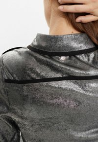 Top in tessuto grigio metallico con texture morbida; presenta un bordo colorato nero sul colletto e un bottone bianco rotondo sul retro.