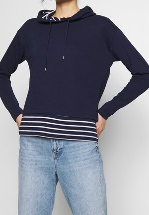Person iført navyblå hættetrøje over marineblå og hvide striber skjorte med lyseblå jeans, hænderne hvilende på hofterne.