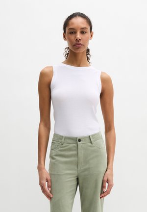 Femme aux cheveux bouclés attachés portant un haut blanc sans manches et un pantalon taille haute vert clair, sur un fond uni.