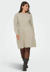 Vestido de punto en beige claro, con mangas largas acanaladas y un torso ajustado, que fluye hacia una falda acampanada; combinado con botas negras hasta la rodilla.