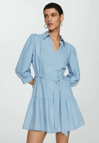 Robe en denim bleu clair avec un décolleté en V, des manches bouffantes et une taille ceinturée. Elle présente une jupe évasée et une texture lisse.