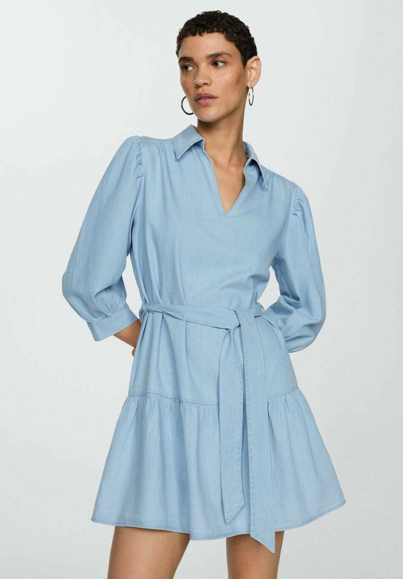 Robe en denim bleu clair avec un décolleté en V, des manches bouffantes et une taille ceinturée. Elle présente une jupe évasée et une texture lisse.