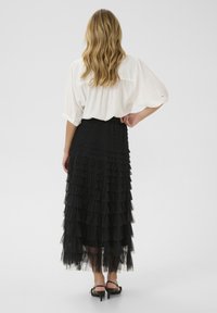 Black tiered midi skirt made of tulle with ruffled layers; worn with a loose white blouse. Strappy, black heeled sandals visible.