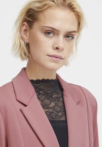 Blazer aus zartem pinken Stoff mit Revers, über einem schwarzen Spitzentop mit hohem Kragen getragen. Geschmeidige Textur, taillierte Passform und detailliertes Spitzenmuster.