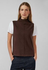 Braunes, ärmelloses Strickoberteil mit hohem, geripptem Kragen. Nahtdetail entlang der Vorderseite, darüber eine weiße Kurzarmbluse getragen. Lockerer Schnitt.