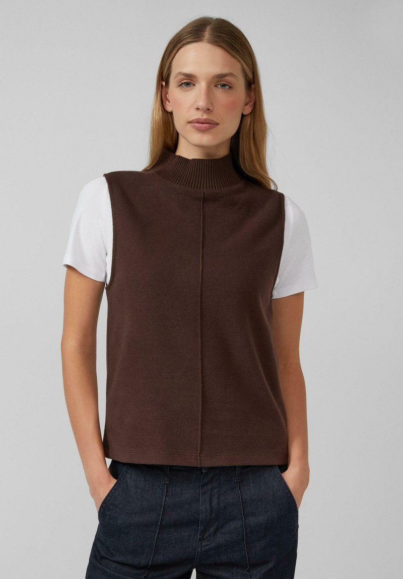 Braunes, ärmelloses Strickoberteil mit hohem, geripptem Kragen. Nahtdetail entlang der Vorderseite, darüber eine weiße Kurzarmbluse getragen. Lockerer Schnitt.