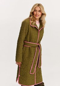 Prošívaný olivovo zelený kabát s ružovými prvkami, zapínaním na gombíky, opaskom v páse a vzorovanou textúrou. Dlhé rukávy a dizajn dĺžky po kolená.