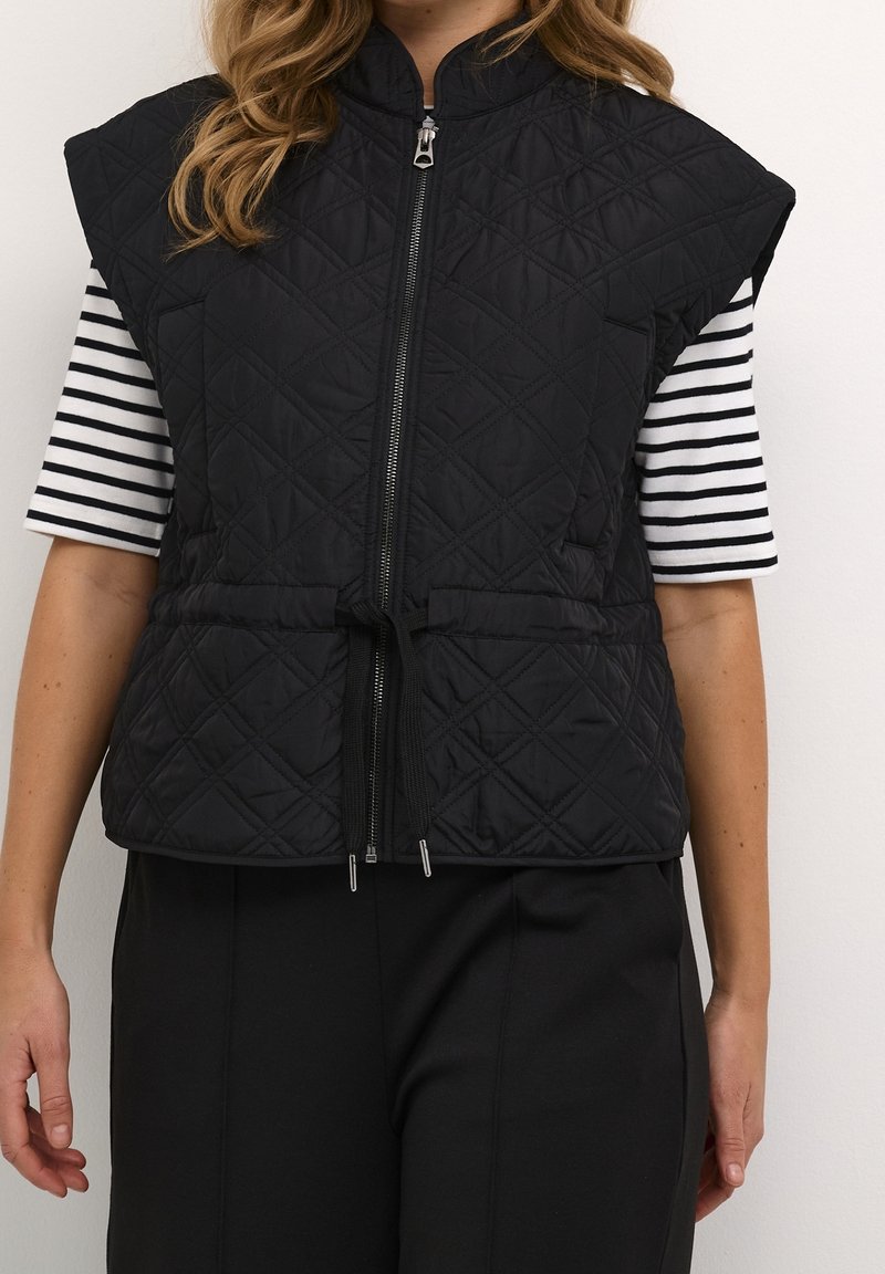 Schwarze gesteppte Weste mit hohem Kragen, durchgehendem Reißverschluss und verstellbarem Kordelzug in der Taille. Begleitet von einem schwarz-weißen gestreiften Hemd.