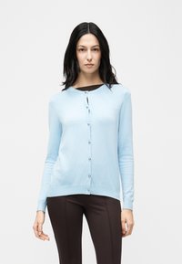 Γυναίκα με μακριά σκούρα μαλλιά, φορούσα ανοιχτό μπλε ζακέτα με κουμπιά πάνω από σκούρο τοπ και στενά σκούρα παντελόνια, στέκεται μπροστά σε απλό λευκό φόντο.