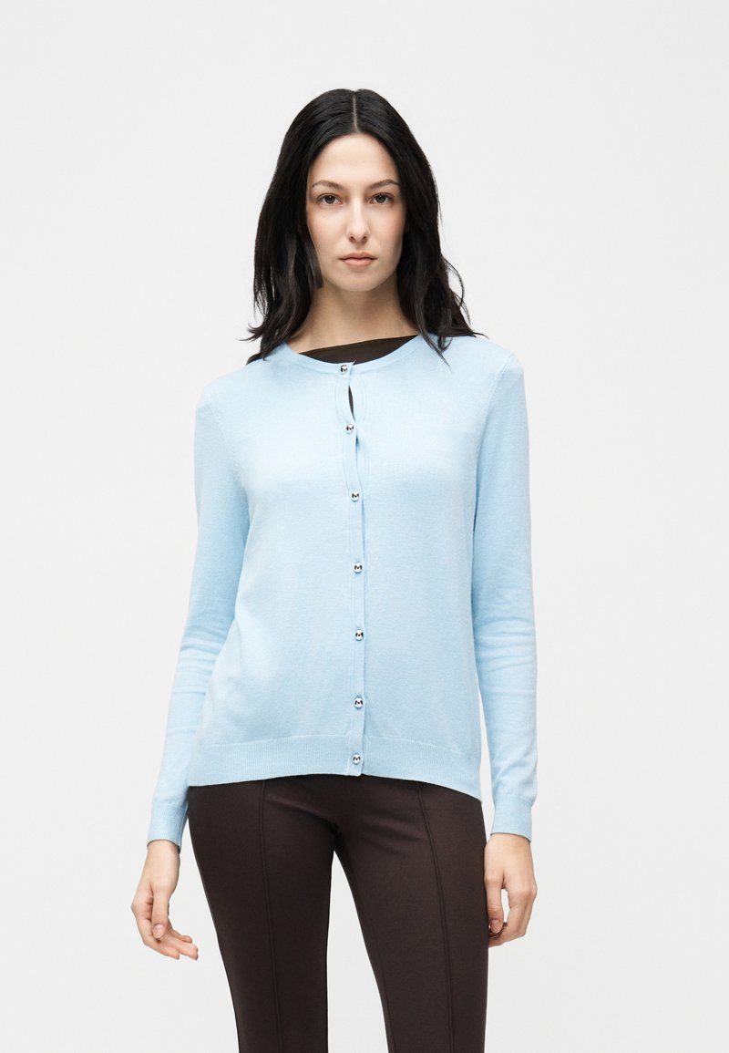 Γυναίκα με μακριά σκούρα μαλλιά, φορούσα ανοιχτό μπλε ζακέτα με κουμπιά πάνω από σκούρο τοπ και στενά σκούρα παντελόνια, στέκεται μπροστά σε απλό λευκό φόντο.