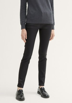 Černé slim-fit kalhoty s hladkou texturou, spárované se šedým pleteným svetrem a černými šněrovacími botami. Viditelný detail s nastavitelným pasem.