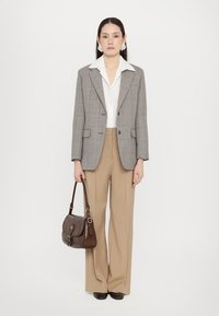 Blazer gri în carouri peste o cămașă albă, asortat cu pantaloni largi bej. Geantă din piele maro cu clapetă curbată și mânere de umăr.