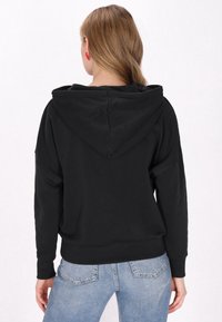 Sudadera con capucha negra de corte holgado y puños acanalados, con una textura de tejido suave. Lleva puesta unos jeans azul claro.