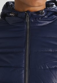 Granatowa kurtka puchowa z wysokim kołnierzem, widocznym zamkiem błyskawicznym oraz pikowaną teksturą. Posiada metalowe elementy i lekkie, błyszczące wykończenie.