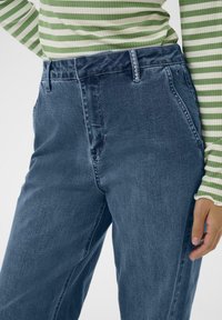 Oseba v modrih kavbojkah in zeleni in beli črtasti dolgimi rokavi, roka počiva ob njeni strani.