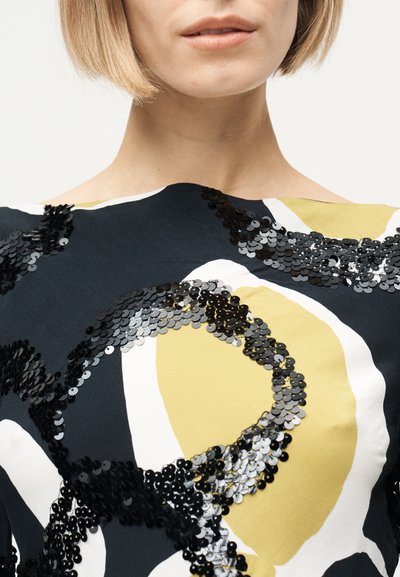 Γυναίκα με κοντά ξανθά μαλλιά που φοράει μπλούζα σε μαύρο, λευκό και κίτρινο χρώμα, διακοσμημένη με μαύρα και ασημένια παγιετένια κυκλικά σχέδια.
