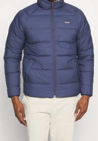 Marinblå isolerad jacka med hög krage, dragkedja framtill och sidofickor. Har quiltade mönster och elastiska ärmslut. Logotyp på bröstet.