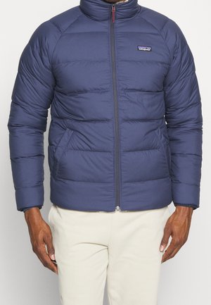 Chaqueta aislante de color azul marino con cuello alto, cierre delantero con cremallera y bolsillos laterales. Presenta un patrón acolchado y puños elásticos. Logo de la marca en el pecho.