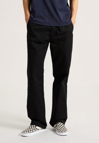 Black pants made of cotton with a straight leg design. Worn with checkerboard slip-on shoes. Simple, functional construction.