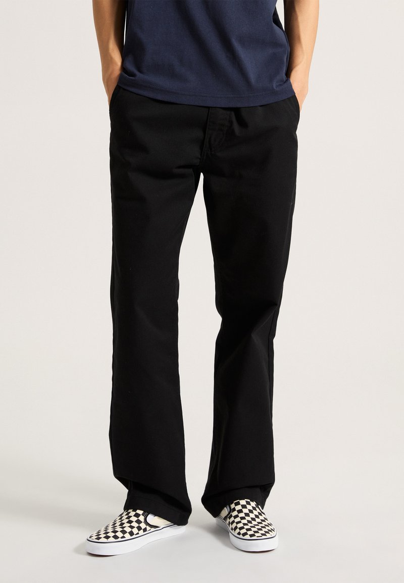 Black pants made of cotton with a straight leg design. Worn with checkerboard slip-on shoes. Simple, functional construction.