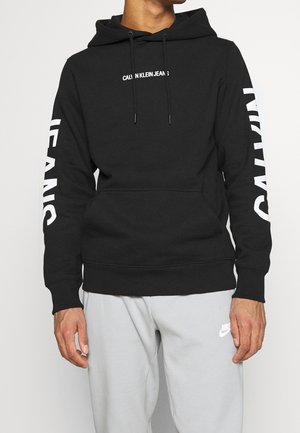 Personne portant un sweat à capuche Calvin Klein noir avec un logo blanc sur la poitrine et les manches, associé à un pantalon de survêtement Nike gris clair.