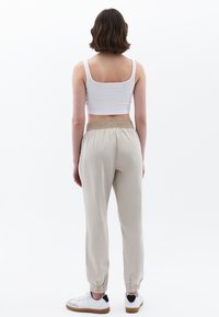 Beige tapered broek met elastische tailleband, licht glanzende textuur, boorden bij de enkels, gecombineerd met een witte crop top en witte sneakers.