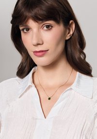 Złota naszyjnik z owalnym zielonym kamieniem szlachetnym i małym białym akcentem, noszony na teksturowanej białej bluzce.