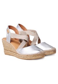 Sandalias de cuña de cuero plateado con tiras cruzadas beige, suela envuelta en yute texturizado y frente plano. Detalles de diseño de punta redondeada.