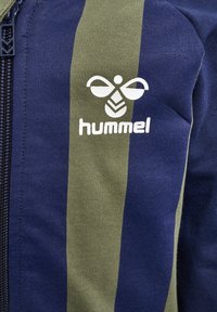 Navy og olivenstribet lynlåsjakke lavet af blødt materiale, med et hvidt logo og struktureret strikmønster.