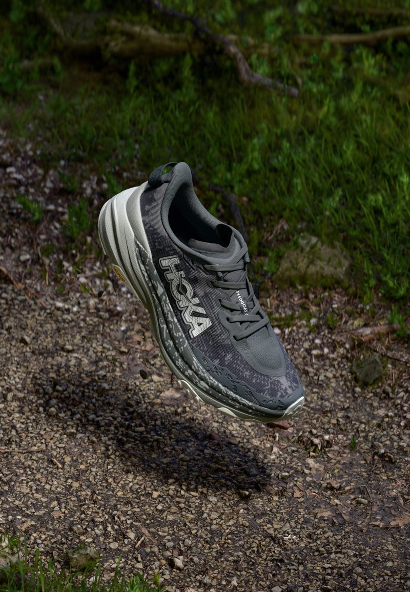 Hoka running shoe in grey with a textured mesh upper, cushioned midsole, distinct tread pattern, and white logo detail.