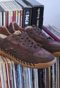 Zapatillas de ante marrón con suelas de goma descansan en una estantería llena de discos de vinilo y libros, incluyendo títulos de Bach y Acercado.