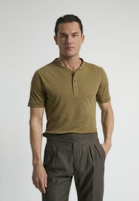 Camisa henley de manga corta en verde oliva con tres botones, combinada con pantalones marrones de cintura alta y plisados. Textura suave, diseño ajustado.