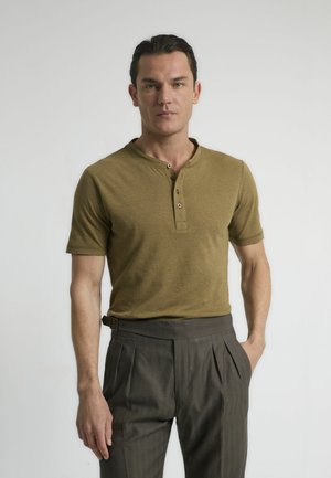Camisa henley de manga corta en verde oliva con tres botones, combinada con pantalones marrones de cintura alta y plisados. Textura suave, diseño ajustado.
