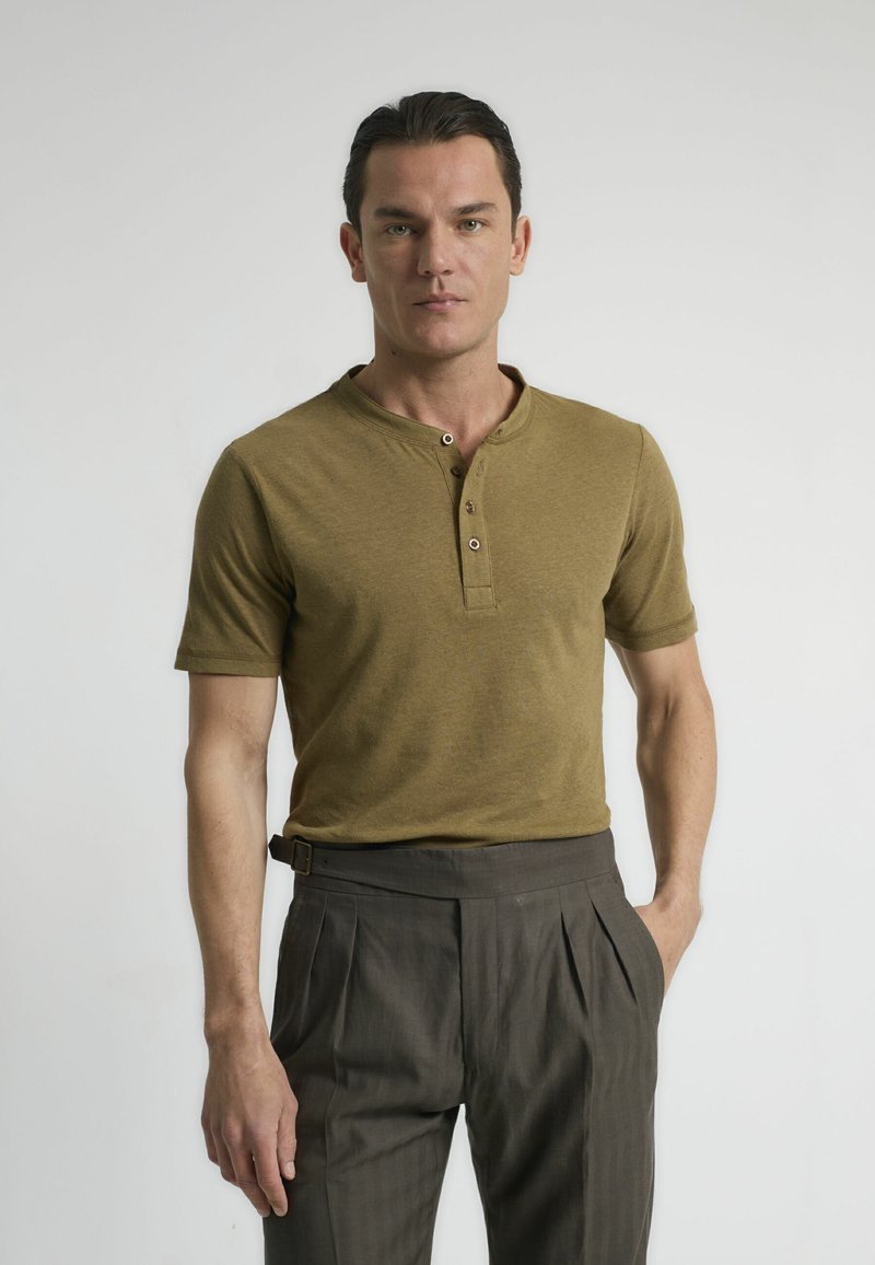 Camisa henley de manga corta en verde oliva con tres botones, combinada con pantalones marrones de cintura alta y plisados. Textura suave, diseño ajustado.