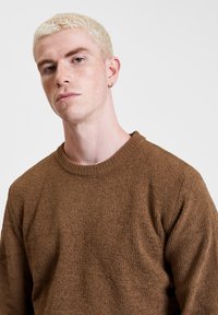 Brązowy dzianinowy sweter o teksturowanym wykończeniu, okrągłym dekolcie i ściągaczach na rękawach, noszony przez modelkę z krótkimi blond włosami.