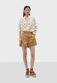 White shirt with red polka dots, two front pockets, paired with brown high-waisted shorts and brown strappy wedge sandals.