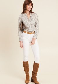 Lichtgekleurde blouse met bladenpatroon, witte skinny jeans, bruin suède kniehoge laarzen en een tan-kleurige riem met een ronde gesp.