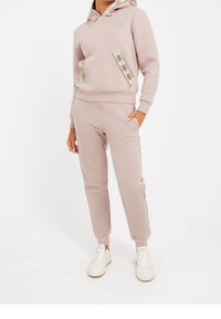 Sudadera con capucha de color rosa claro con ribete estampado y bolsillo tipo canguro, combinada con pantalones jogger a juego con cremalleras laterales y detalles con logo.