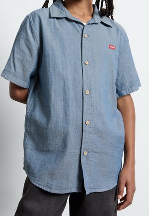 Dziecko ubrane w niebieską koszulę z krótkim rękawem zapinaną na guziki, z czerwonym logo Levi’s na piersi i ciemne spodnie na jednolitym tle.