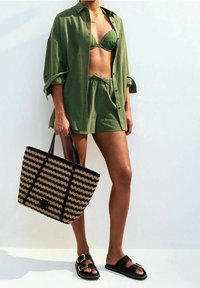 Camisa de tejido verde, bikini y pantalones cortos. Tote con patrón natural y negro, sandalias negras con acentos de hebilla. Texturas suaves y estilo relajado.