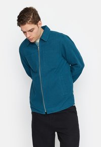 Teal zip-up jas met een kraag en twee borstzakken, gemaakt van een gestructureerde stof. Slim fit, gecombineerd met zwarte broeken.