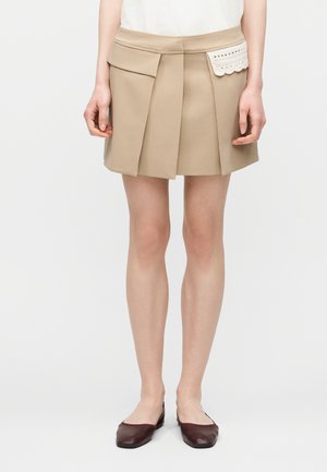 Μπεζ πλισέ μίνι φούστα με μεγάλο καπάκι τσέπης αριστερά και μικρή τσέπη με δαντέλα δεξιά, συνδυασμένη με καφέ μπαλαρίνες και λευκό τοπ.