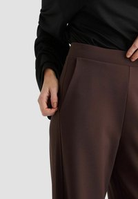 Braune maßgeschneiderte Hose mit Falten, aus einem glatten Stoff. Sie verfügt über Seitentaschen und einen taillierten Bund, der durch ein strukturiertes schwarzes Oberteil ergänzt wird.