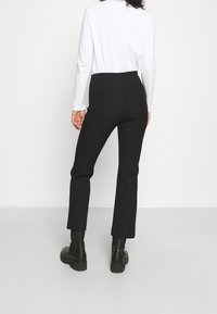 Černé přizpůsobené kalhoty s rovnými nohavicemi, se zadními kapsami, spojené s černými kotníkovými botami na jednobarevném pozadí.