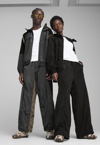 Twee modellen dragen oversized, zwart-grijze trainingspakken van glanzend, textuurrijk materiaal, gecombineerd met schoeisel met camouflagepatroon.
