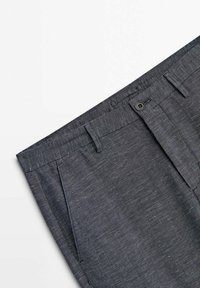 Gros plan d’un pantalon habillé gris foncé montrant la ceinture, les passants, le bouton et la poche latérale en tissu texturé.
