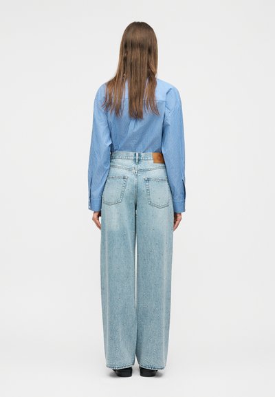 Άτομο με μακριά μαλλιά που φοράει μπλε ριγέ πουκάμισο και τζιν παντελόνι ψηλόμεσο με φαρδύ πόδι σε ανοιχτό μπλε χρώμα με μικρές διακοσμήσεις, στέκεται με την πλάτη προς τα εμπρός.