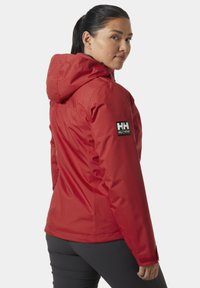 Czerwona wodoodporna kurtka z kapturem, o smukłym designie i regulowanymi mankietami. Posiada naszywkę z logo na lewym rękawie. Gładka tekstura.