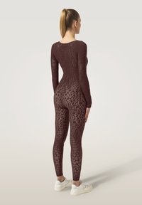 Burgundy långärmad bodysuit med ett strukturerat nätmönster och åtsittande design, kombinerad med vita sneakers, sedd bakifrån.