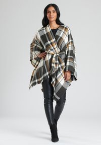 Randig poncho i mjukt tyg i vita, svarta och bruna nyanser, med bälte i midjan och fransade kanter. Bärs över åtsittande svarta byxor.